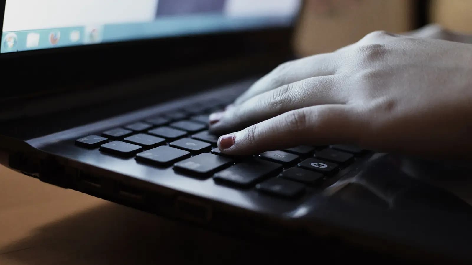 Mão digitando no teclado de um notebook preto em ambiente com pouca luz