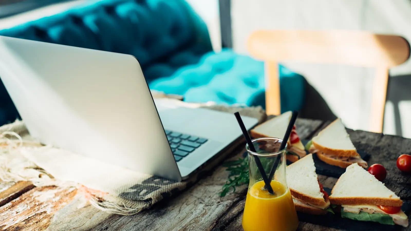 Notebook aberto sobre mesa de madeira em um café, ao lado de sanduíches e copo de suco de laranja