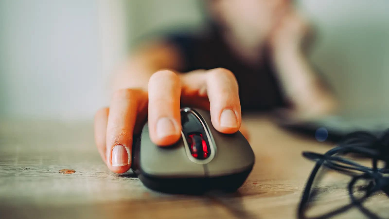 Mão apoiada sobre um mouse de computador prestes a clicar, com pessoa desfocada ao fundo