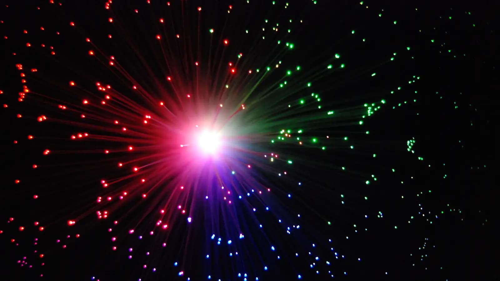 Feixes de fibra óptica iluminados em vermelho, verde e azul irradiando de um ponto de luz no escuro