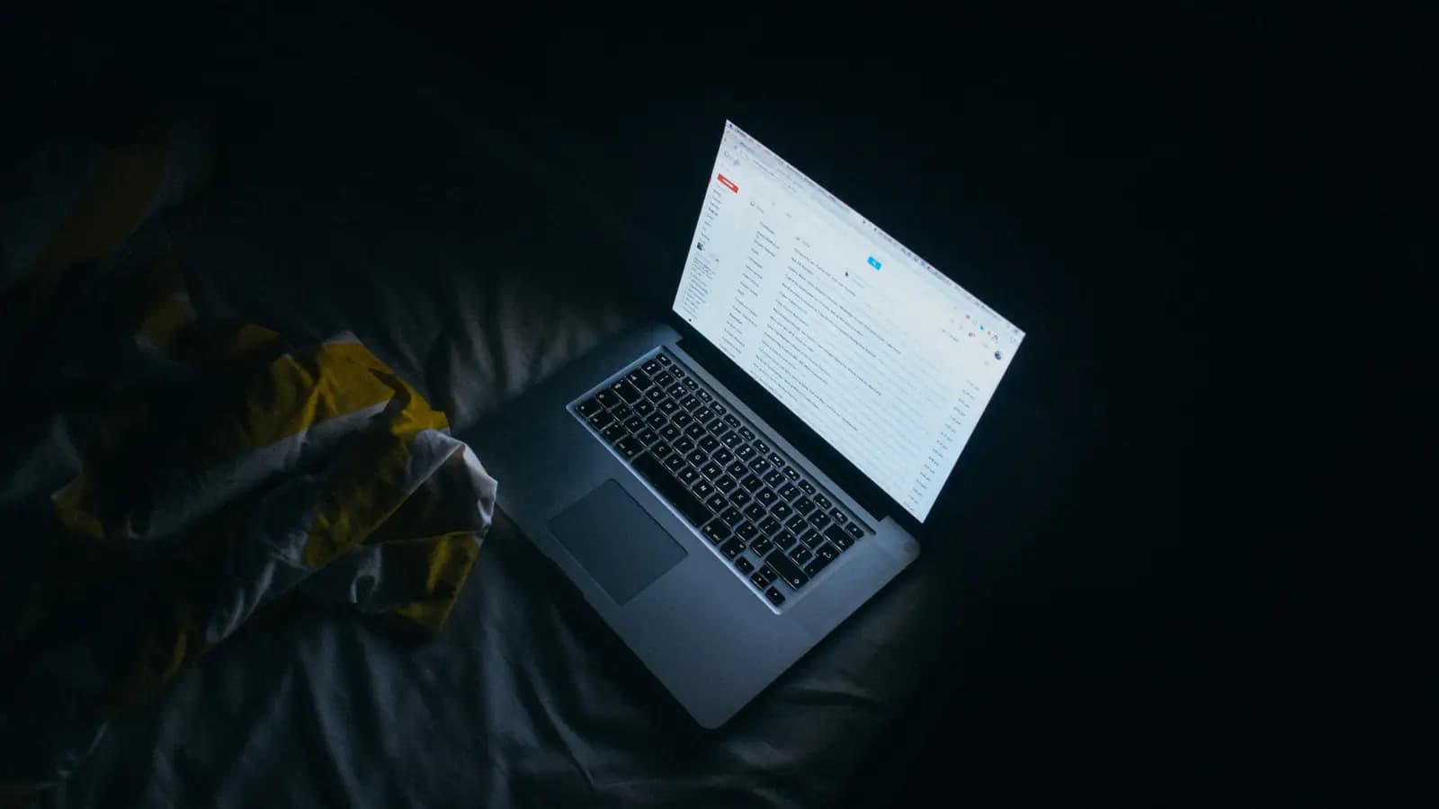 Notebook aberto com a tela acesa iluminando uma cama em um quarto escuro durante a noite
