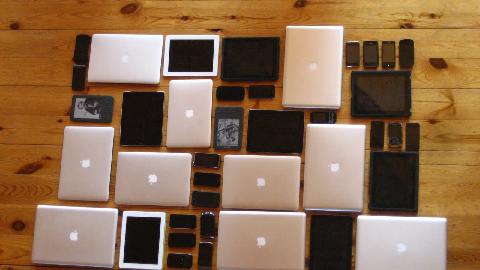 Vários notebooks, tablets, celulares e leitores digitais organizados lado a lado sobre piso de madeira